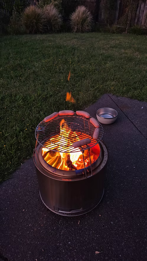 CleanBurn fire pit with hotdogs on a grill in a backyard